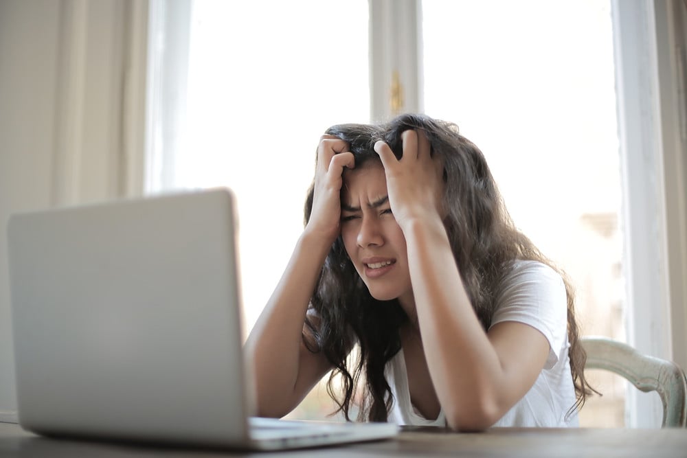 Woman sitting in front of laptop with hands in hair on side of head looking very stressed