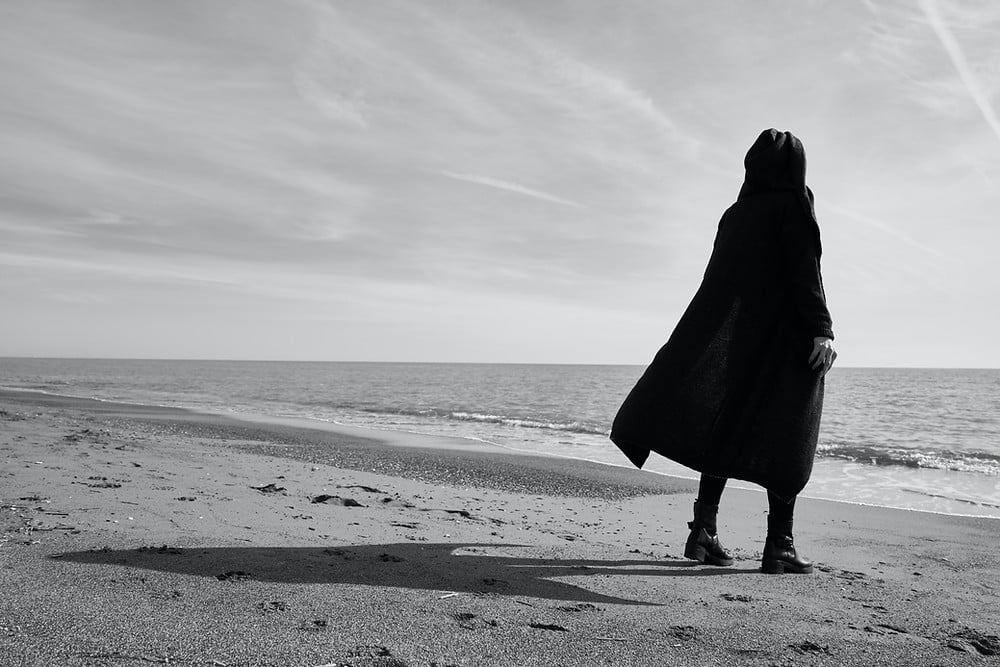Person standing on the beach facing the water dressed all in black with hood up covering their head